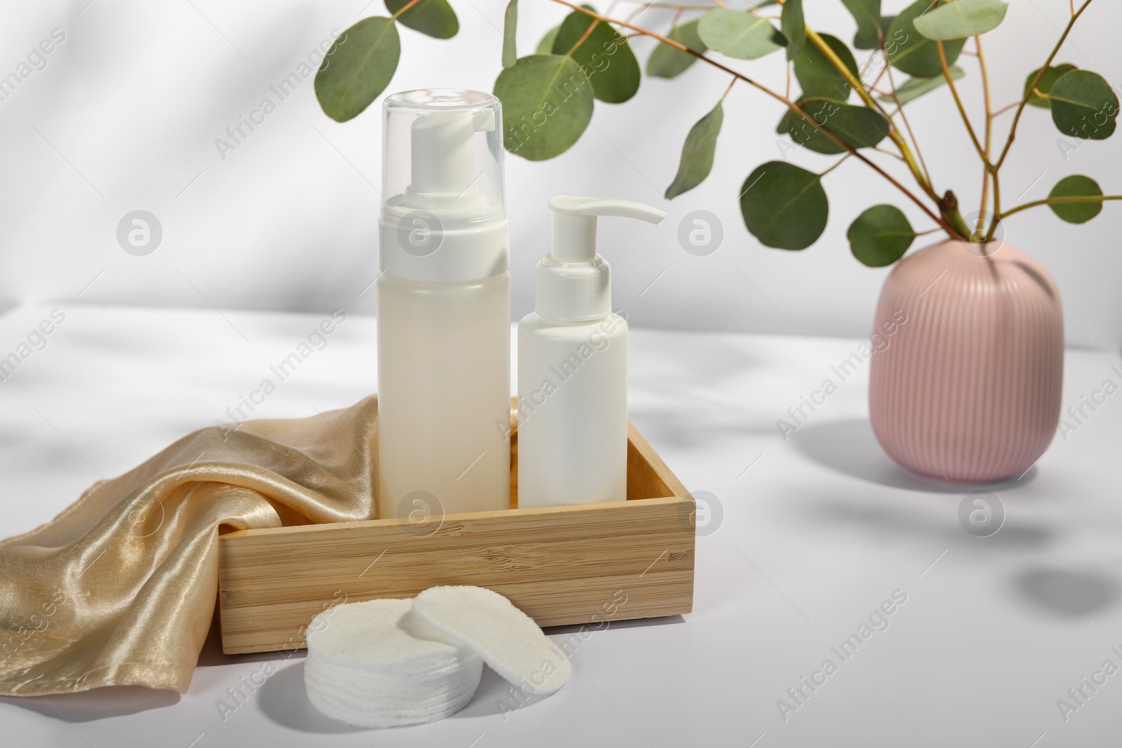 Photo of Bottles of face cleansing products, cotton pads and eucalyptus branches on white table