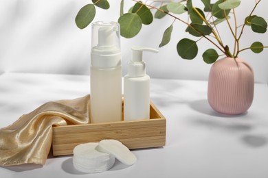 Bottles of face cleansing products, cotton pads and eucalyptus branches on white table