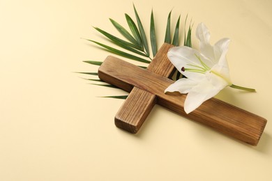 Photo of Wooden cross, lily flower and palm leaf on pale yellow background, space for text. Easter attributes