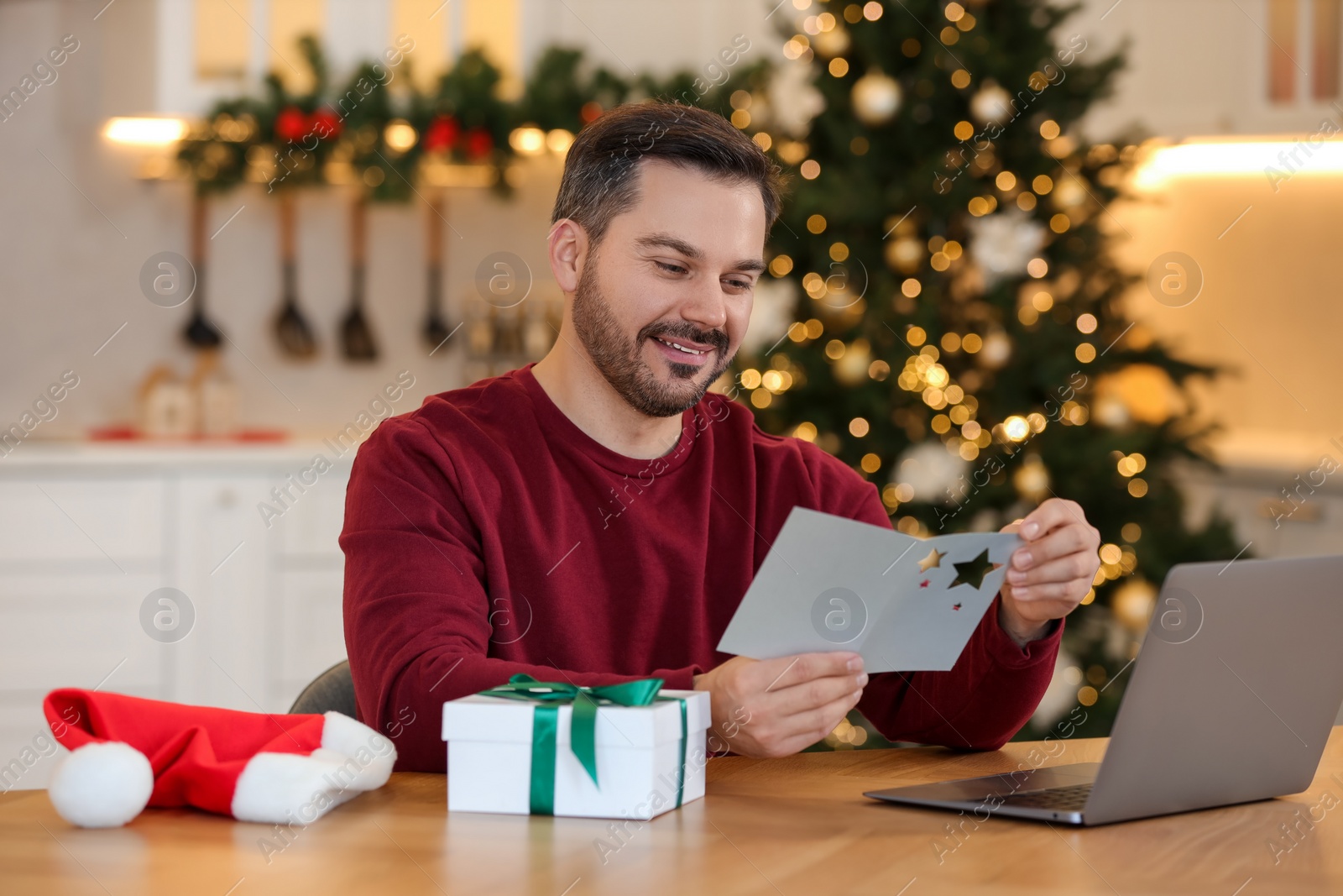 Photo of Celebrating Christmas online with exchanged by mail presents. Happy man reading greeting card during video call on laptop at home