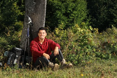 Photo of Hiker with backpack and trekking poles sitting near tree on grass. Rest stop