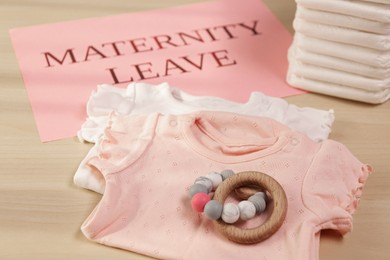 Baby clothes, toy, diapers and note with text Maternity Leave on wooden table, closeup
