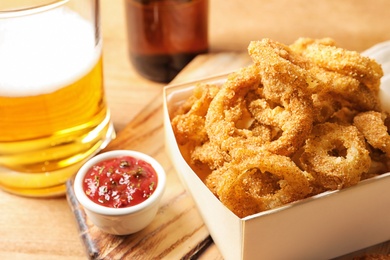 Cardboard box with crunchy fried onion rings and tomato sauce on wooden board, closeup