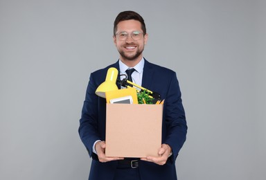 Happy unemployed man with box of personal office belongings on grey background