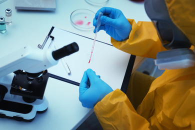 Photo of Scientist in chemical protective suit dripping blood on microscope slide at table, closeup. Virus research