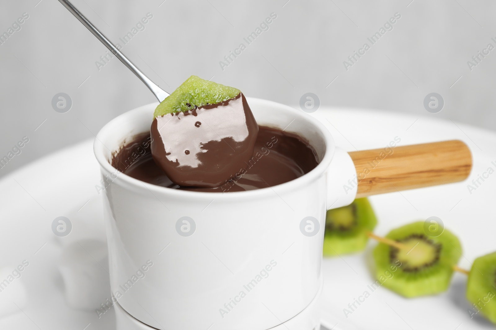 Photo of Dipping kiwi into fondue pot with chocolate on white table