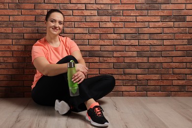 Photo of Happy overweight woman with bottle of water sitting near brick wall indoors, space for text