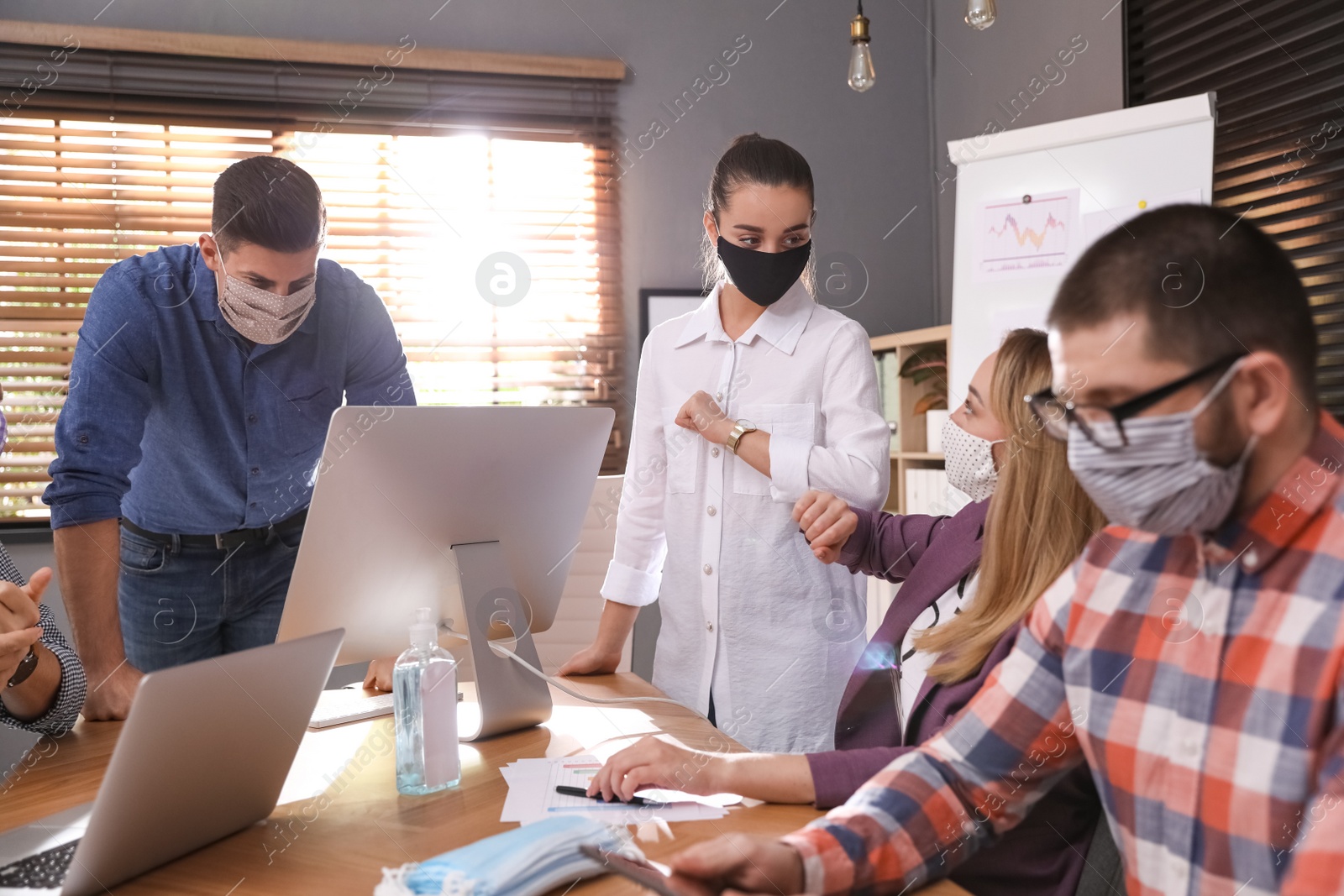 Photo of Coworkers with protective masks making elbow bump in office. Informal greeting during COVID-19 pandemic