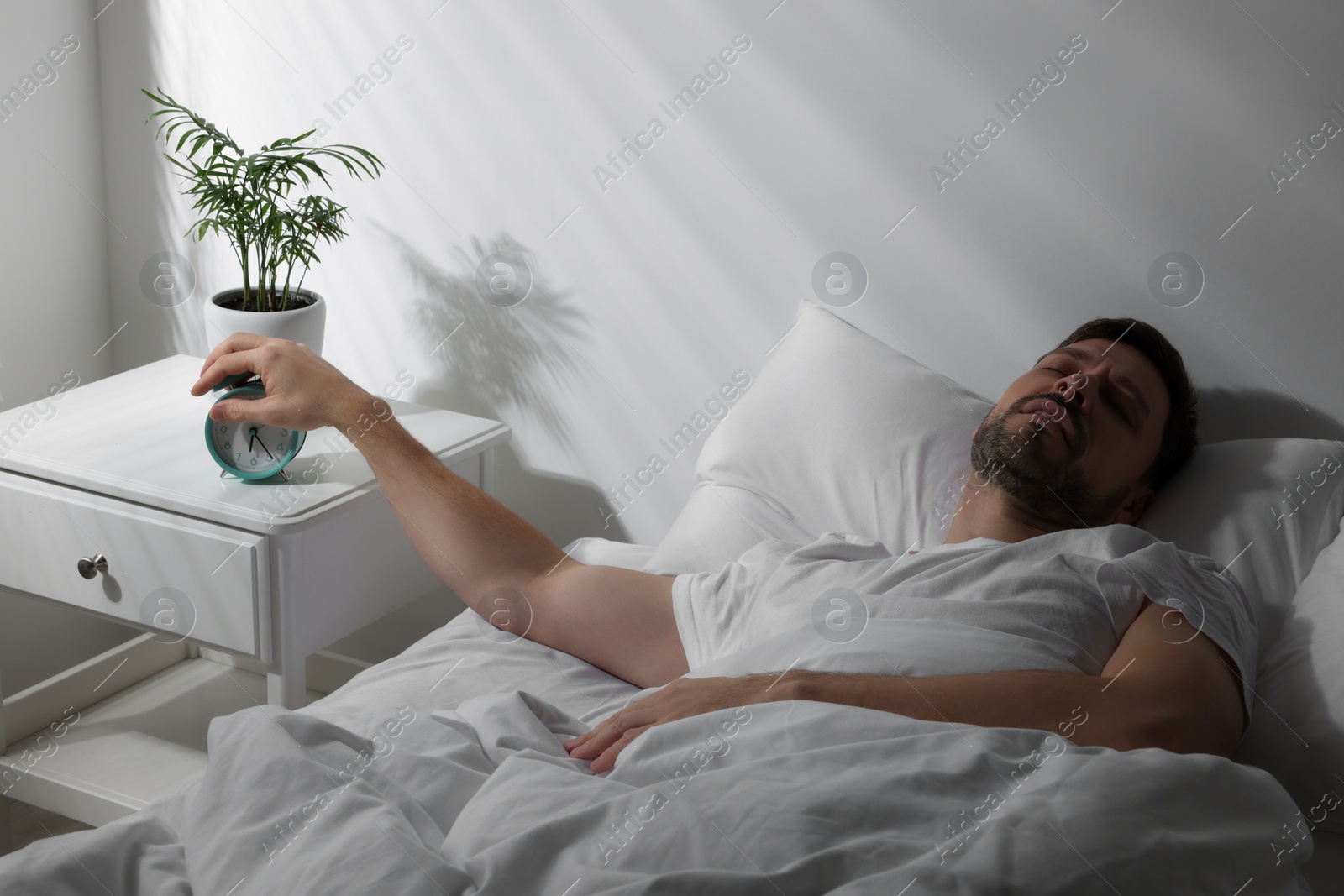 Photo of Sleepy man turning off alarm clock on nightstand in morning