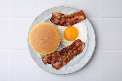 Photo of Plate with tasty pancakes, fried egg and bacon on white tiled table, top view