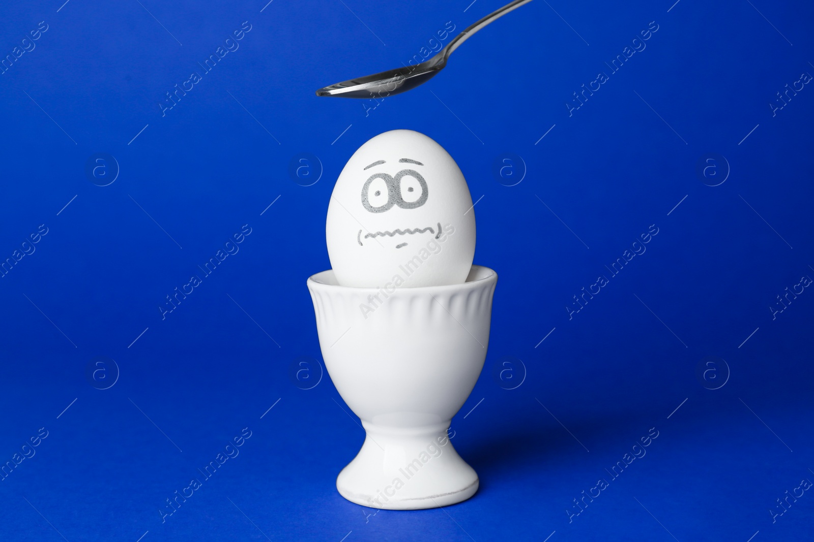 Photo of Spoon over egg with drawn frightened face among others in cup on blue background