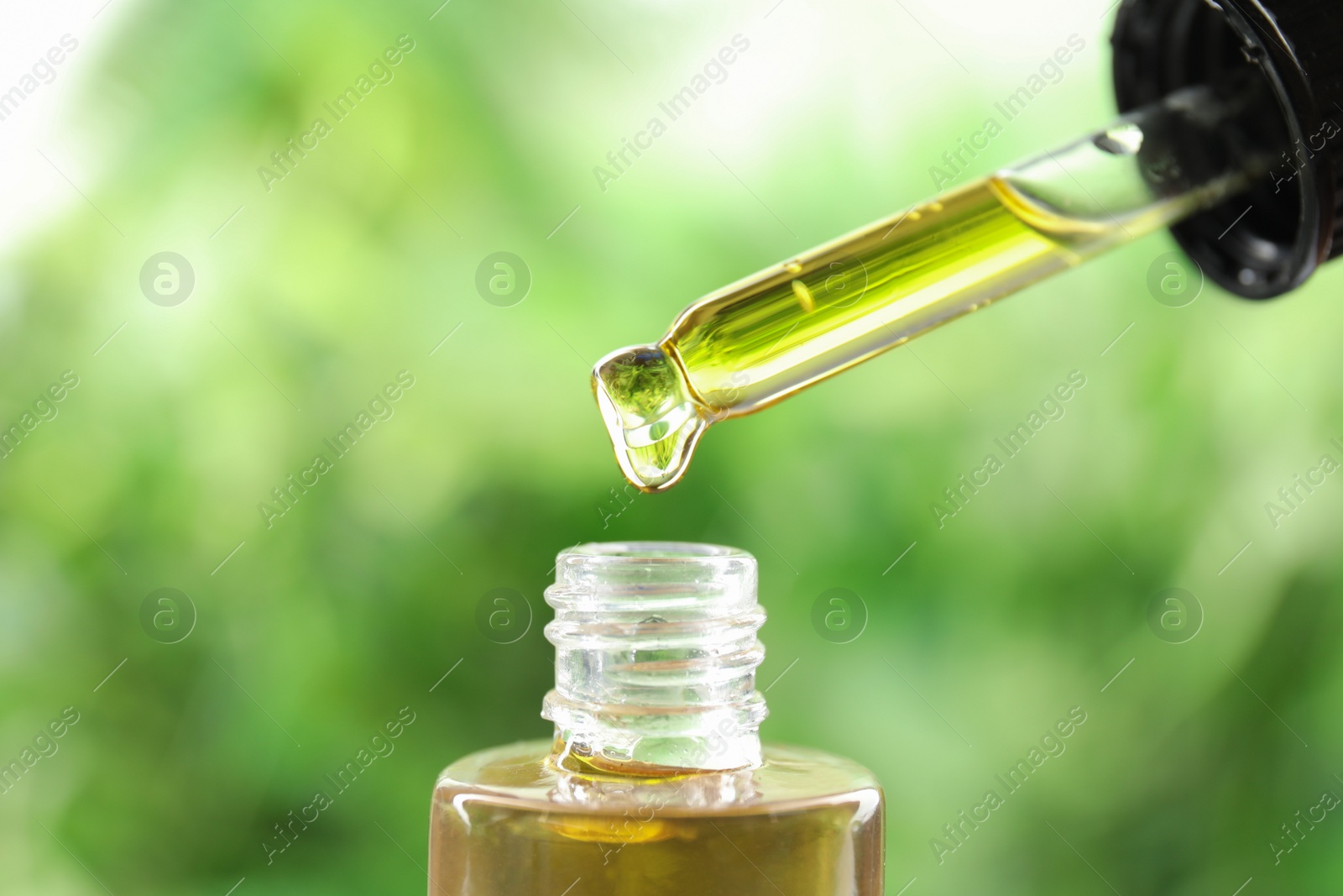 Photo of Dripping essential oil from pipette into glass bottle against blurred green background, closeup