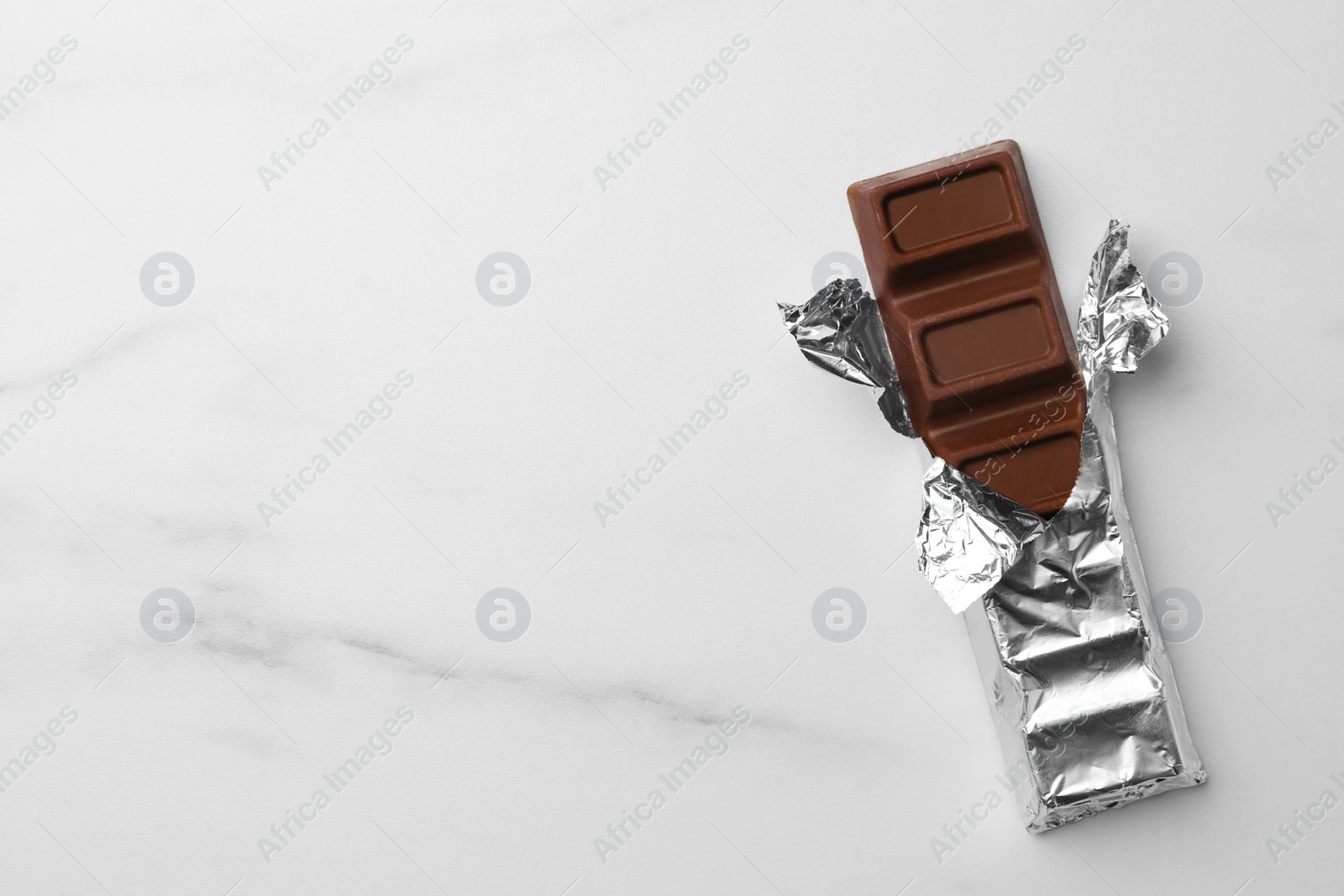 Photo of Tasty chocolate bar wrapped in foil on white table, top view. Space for text