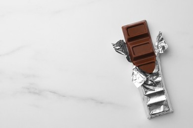 Photo of Tasty chocolate bar wrapped in foil on white table, top view. Space for text