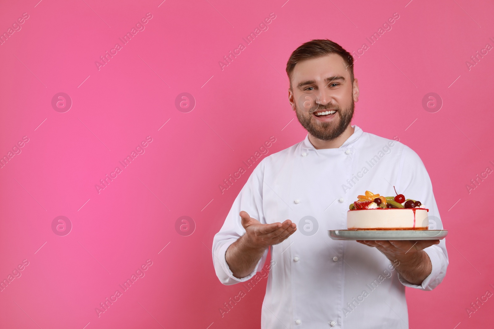 Photo of Happy professional confectioner in uniform holding delicious cake on pink background. Space for text