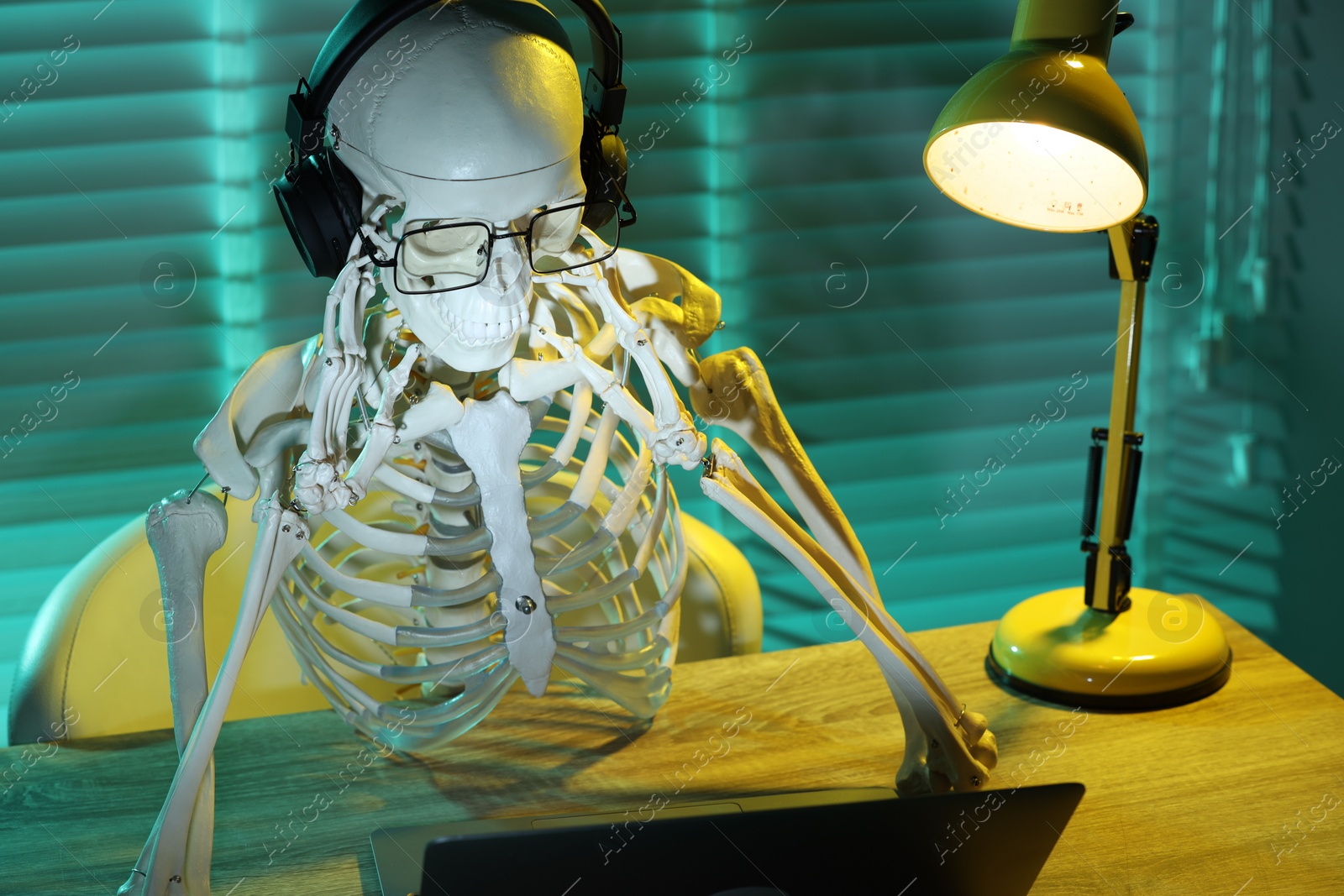 Photo of Waiting concept. Human skeleton sitting at wooden table with laptop and headphones indoors