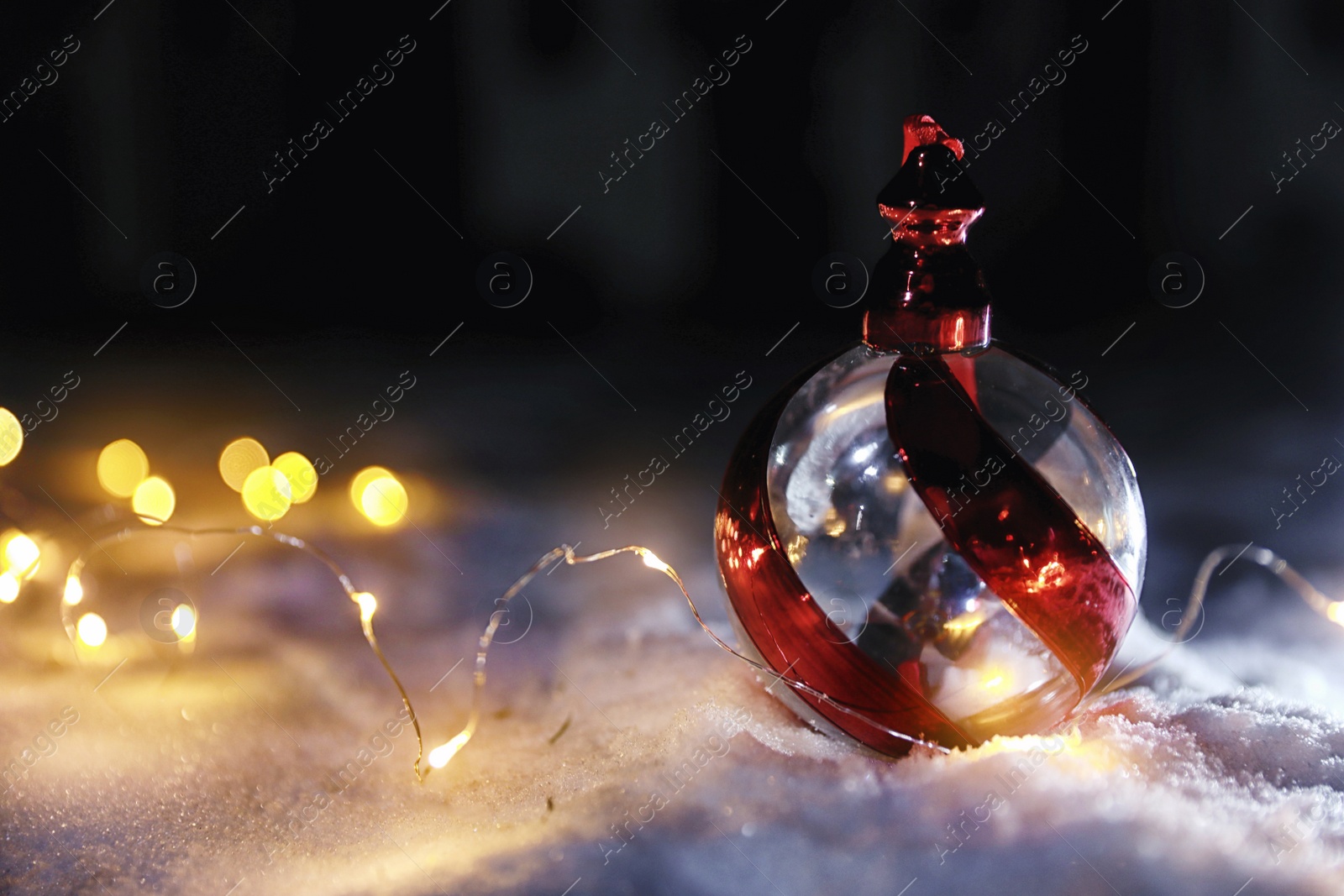 Photo of Christmas ball and fairy lights on white snow outdoors. Space for text