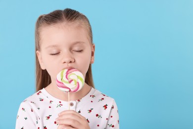 Photo of Cute little girl licking colorful lollipop swirl on light blue background, space for text