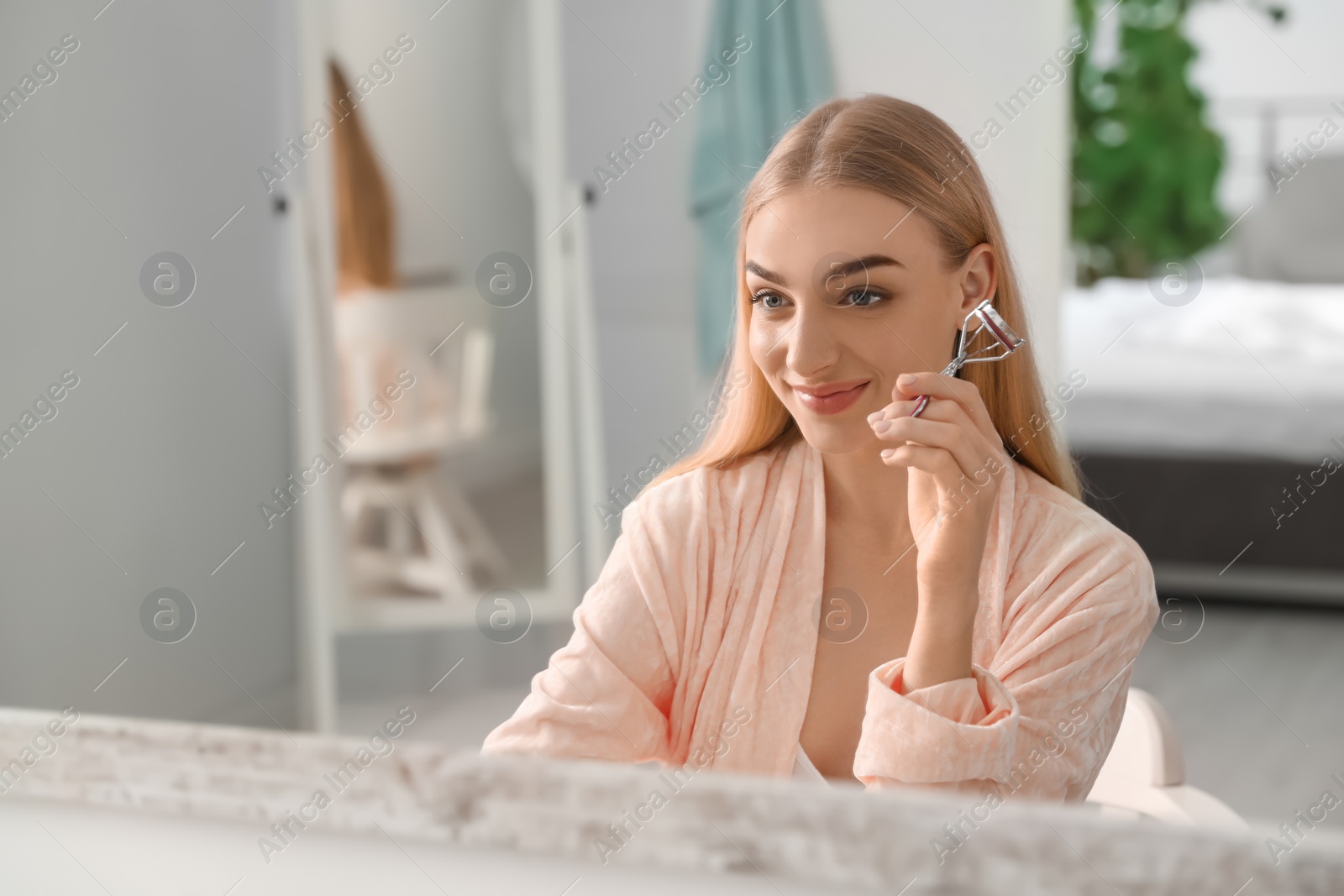 Photo of Young woman curling her eyelashes near mirror indoors