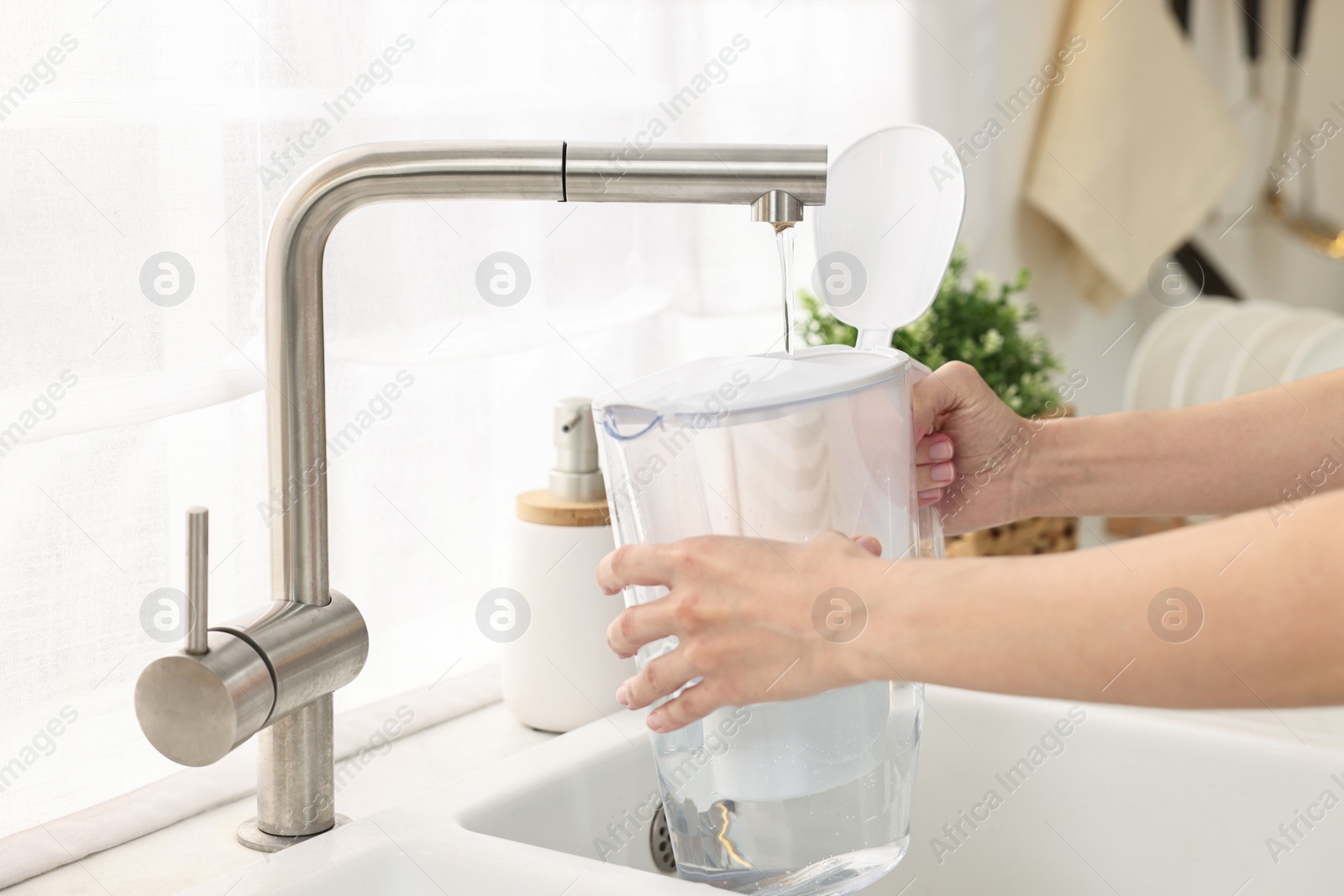 Photo of Woman pouring tap water into filter jug in kitchen, closeup