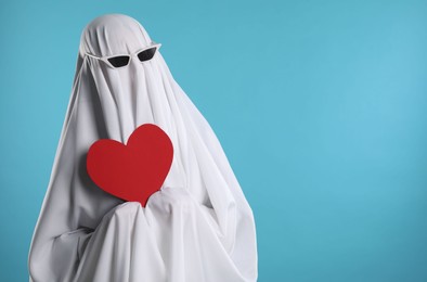 Photo of Cute ghost. Person covered with white sheet in sunglasses holding red heart on light blue background, space for text