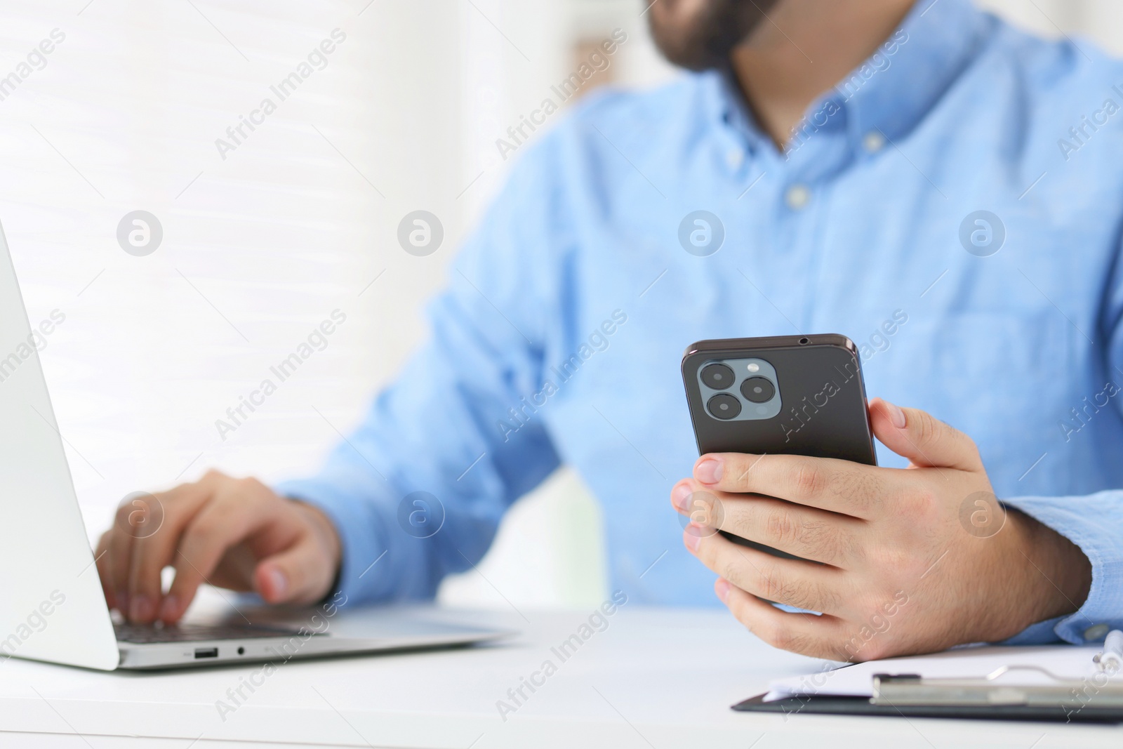 Photo of Young man using smartphone while working with laptop at white table in office, closeup