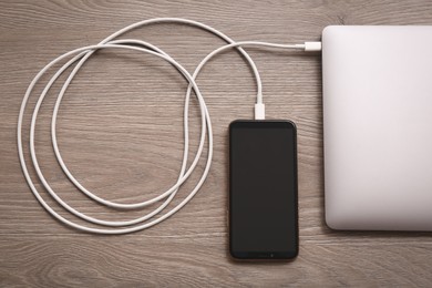 Photo of Smartphone connected with charge cable to laptop on wooden table, flat lay. Space for text