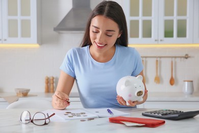 Photo of Young woman with piggy bank counting money at table in kitchen