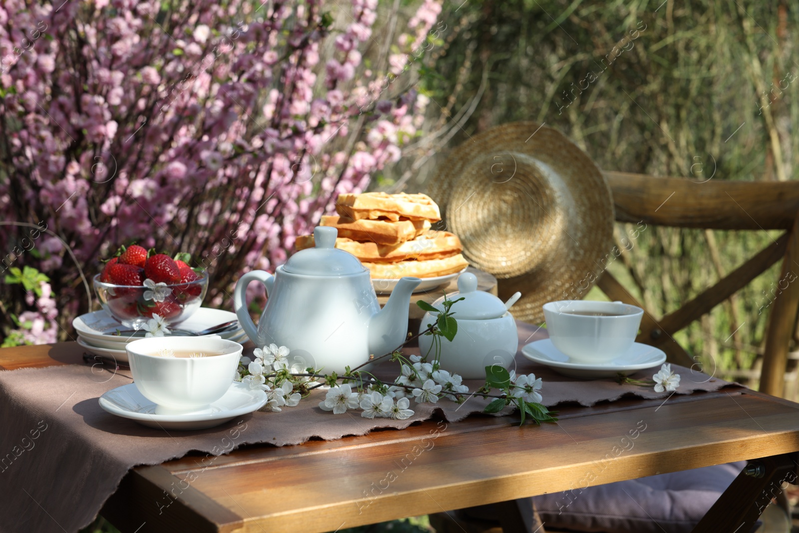 Photo of Beautiful spring flowers, freshly baked waffles and ripe strawberries on table served for tea drinking in garden