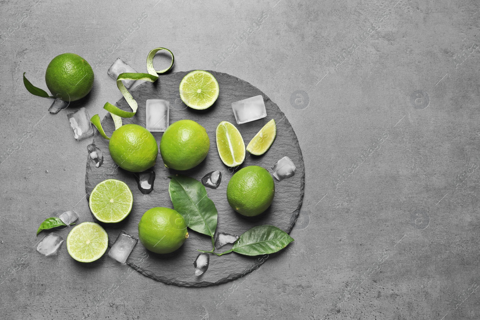 Photo of Slate plate with fresh ripe limes and ice cubes on gray background, top view