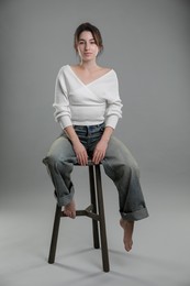 Beautiful young woman sitting on stool against grey background