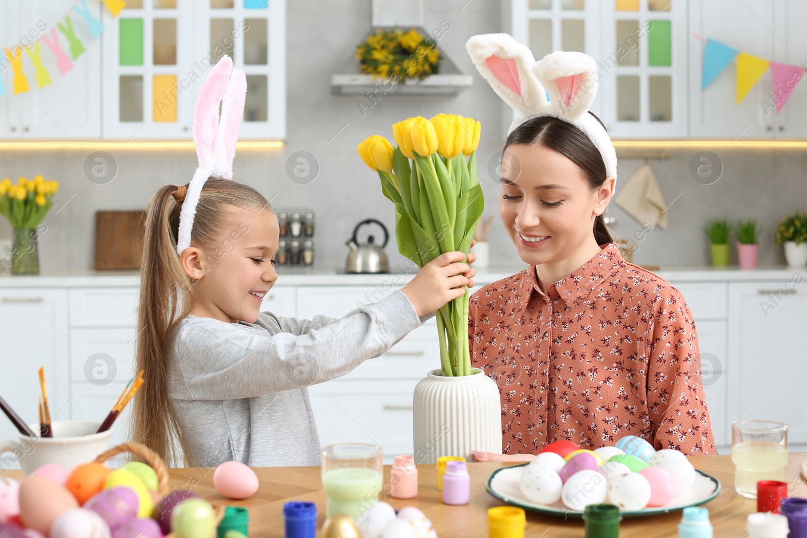 Photo of Mother with her cute daughter putting yellow tulips into vase near beautifully painted Easter eggs at table in kitchen