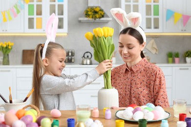 Mother with her cute daughter putting yellow tulips into vase near beautifully painted Easter eggs at table in kitchen