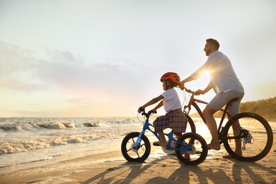 Photo of Happy father with son riding bicycles on sandy beach near sea at sunset