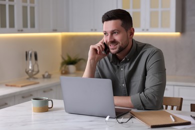 Photo of Happy man talking on phone while working with laptop at white marble table in kitchen