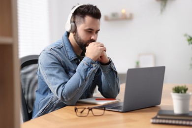 Photo of Young man in headphones watching webinar at table in room