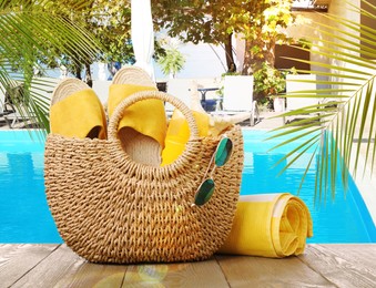 Stylish bag with beach accessories on wooden surface near outdoor swimming pool