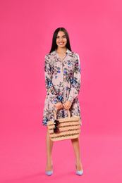 Photo of Young woman wearing floral print dress with straw bag on pink background