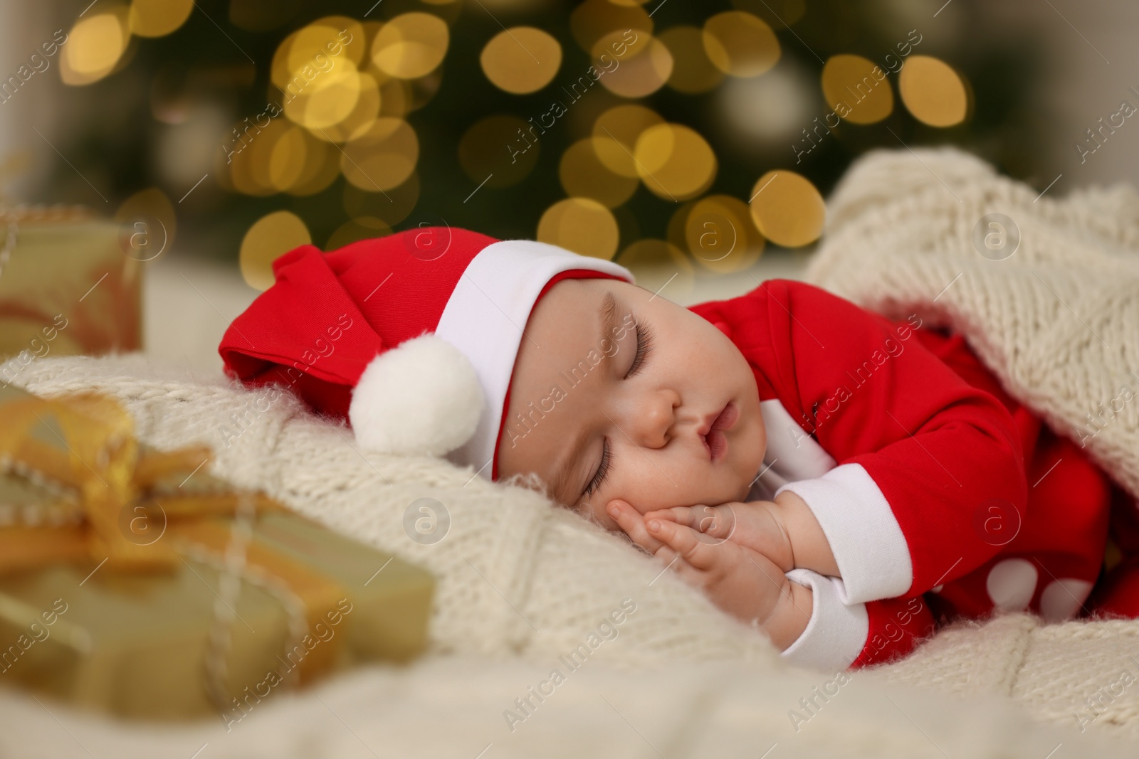 Photo of Cute baby in Christmas costume sleeping on knitted blanket against blurred festive lights. Winter holiday