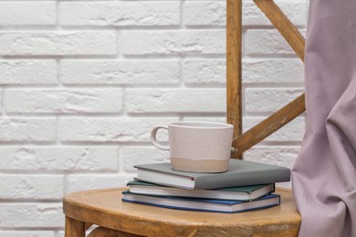 Ceramic cup and stack of books on wooden chair against white brick wall, space for text