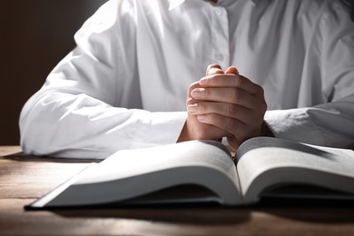 Woman holding hands clasped while praying over Bible at wooden table, closeup