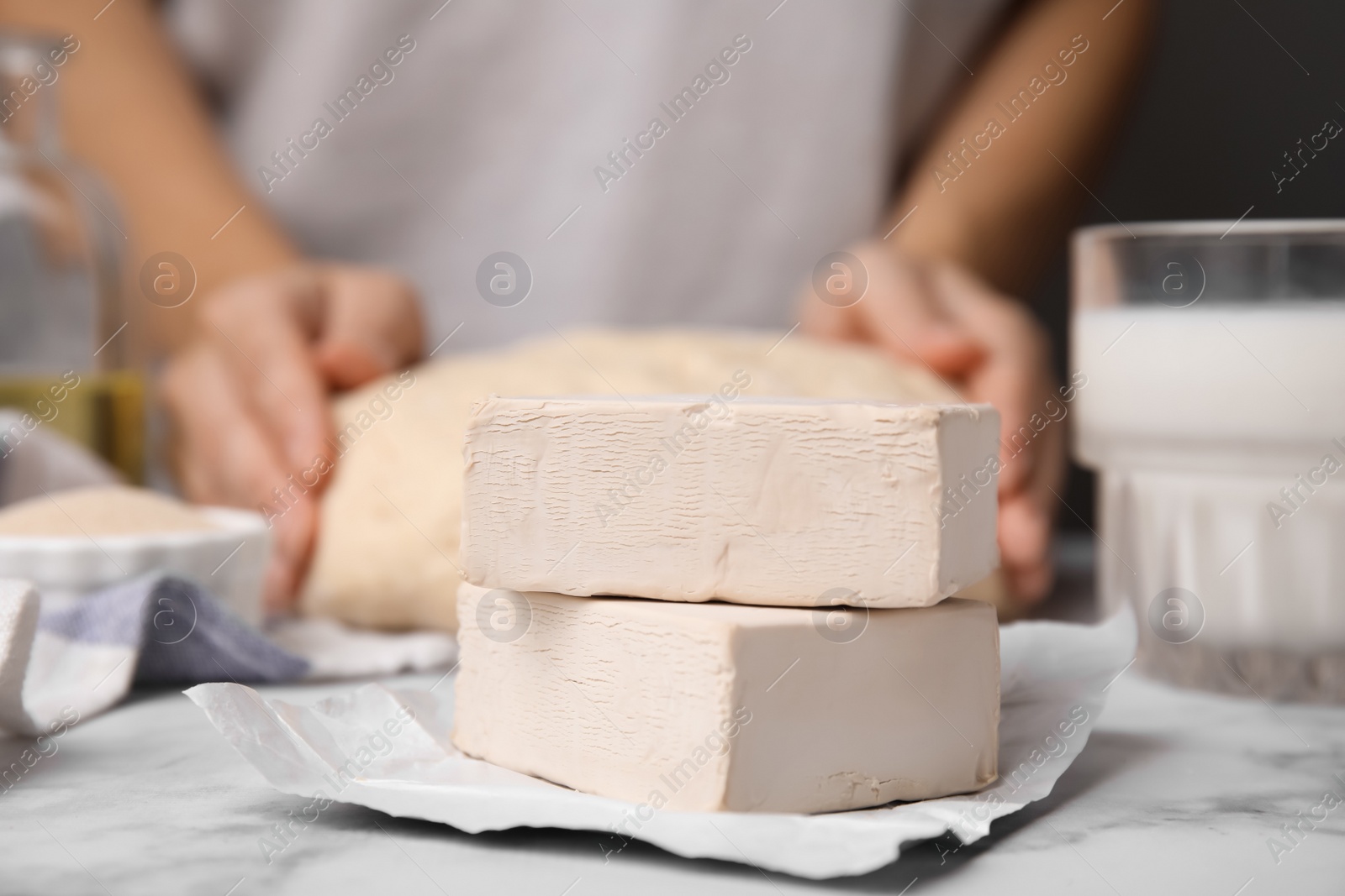 Photo of Woman kneading dough at table, focus on blocks of compressed yeast