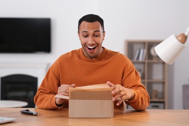 Photo of Happy young man opening parcel at table indoors. Internet shopping