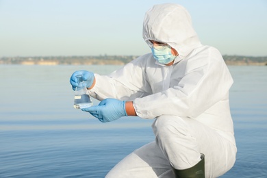 Photo of Scientist in chemical protective suit with conical flask taking sample from river for analysis