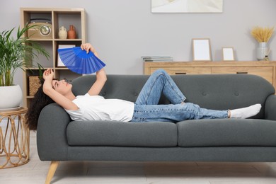 Photo of Young woman waving blue hand fan to cool herself on sofa at home