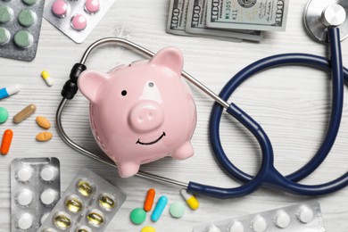 Pale pink ceramic piggy bank, stethoscope, money and pills on white wooden table, flat lay. Medical insurance