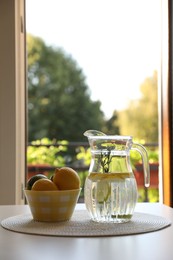 Photo of Jug with refreshing lemon water and citrus fruits in bowl on table indoors