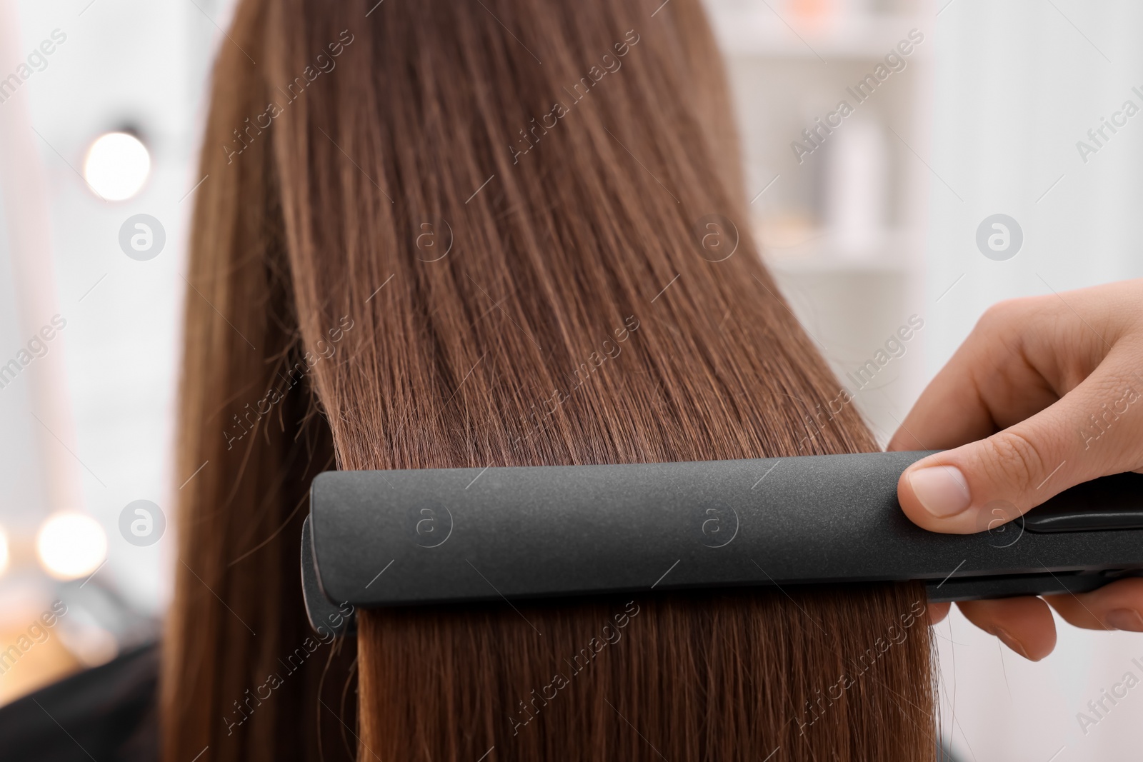 Photo of Hairdresser straightening woman's hair with flat iron indoors, closeup