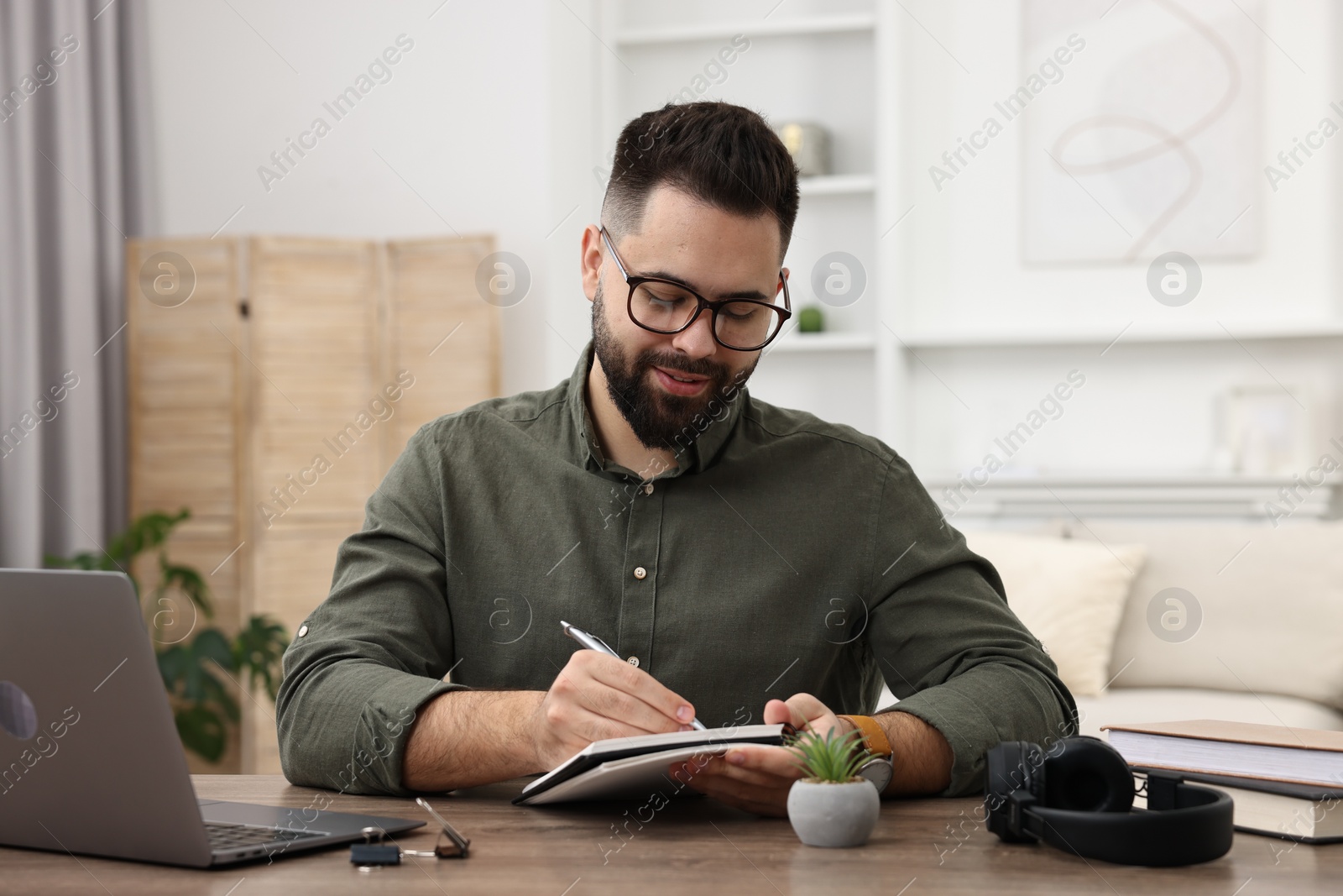 Photo of E-learning. Young man taking notes during online lesson at wooden table indoors