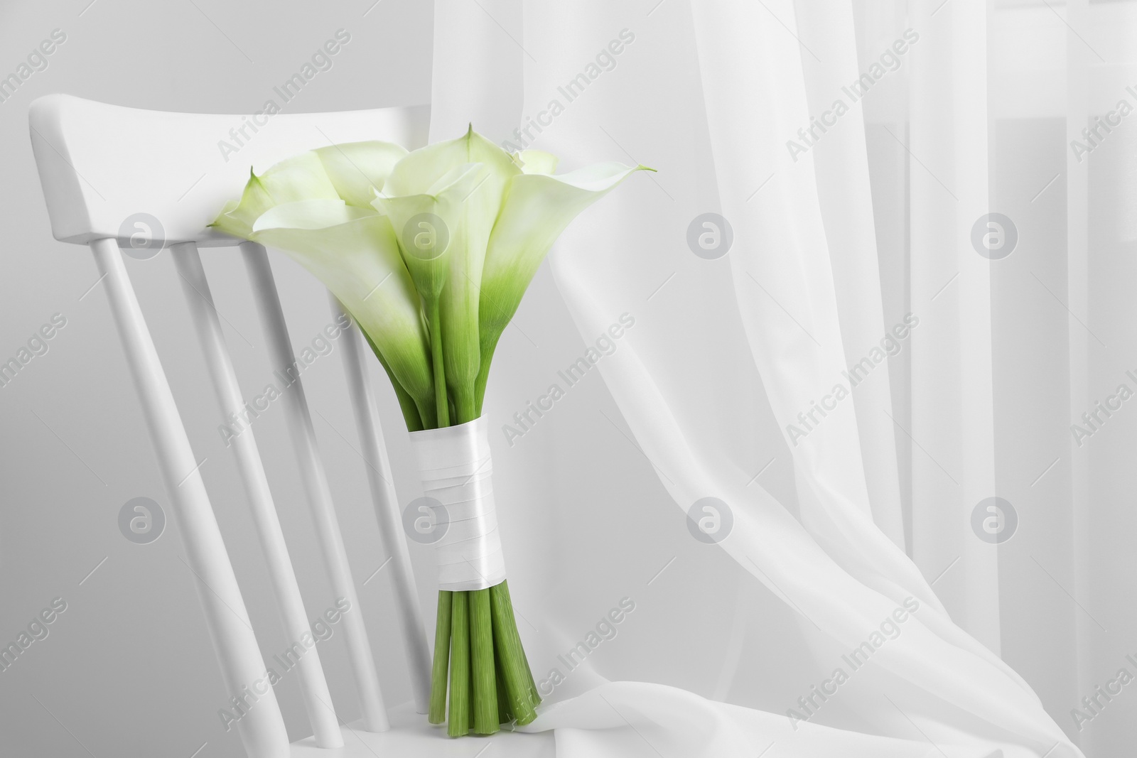 Photo of Beautiful calla lily flowers tied with ribbon on white chair indoors